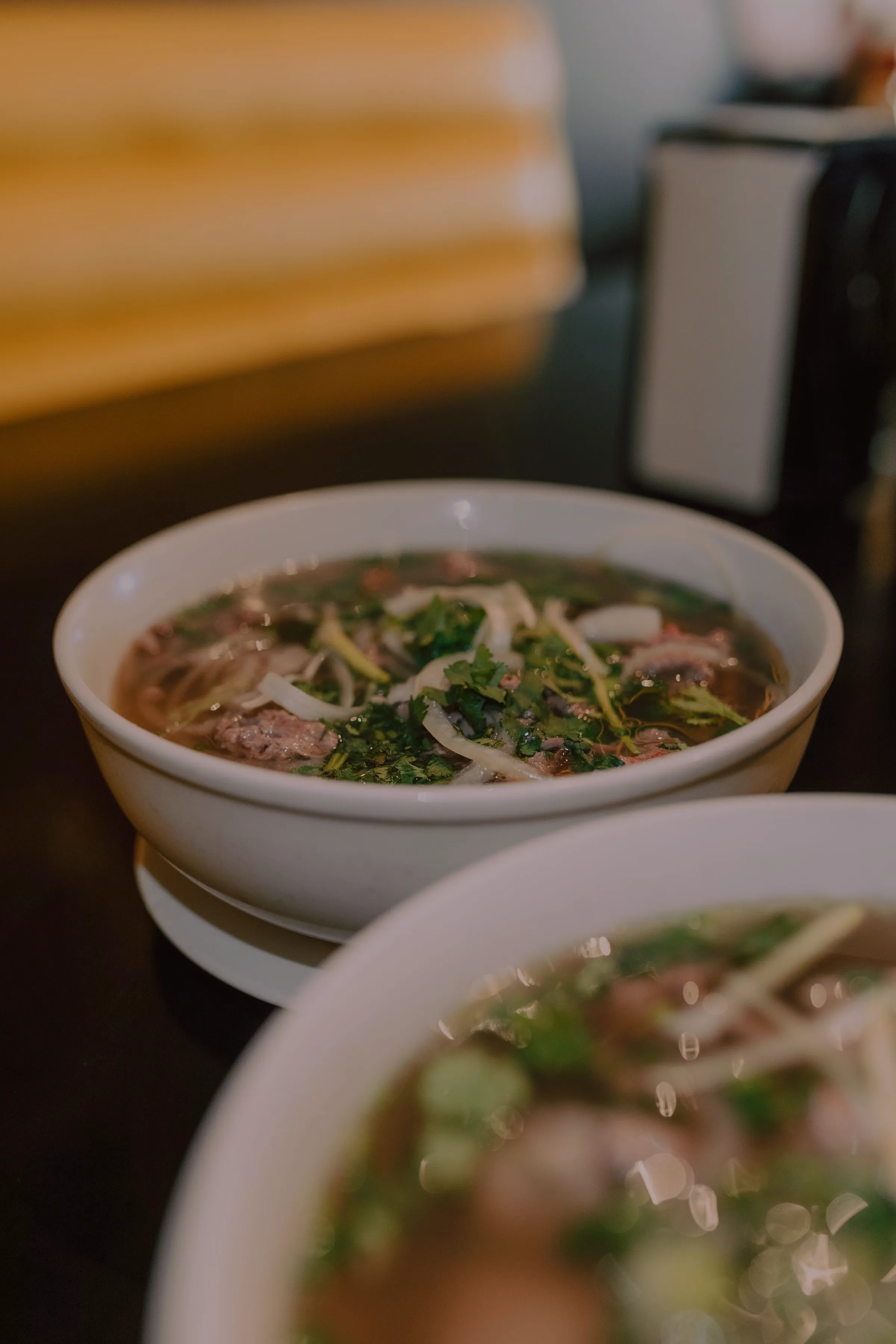 Two bowls of pho on a dark glossy table, the nearer one thrown out of focus in the foreground, with a warm amber banquette glowing softly behind
