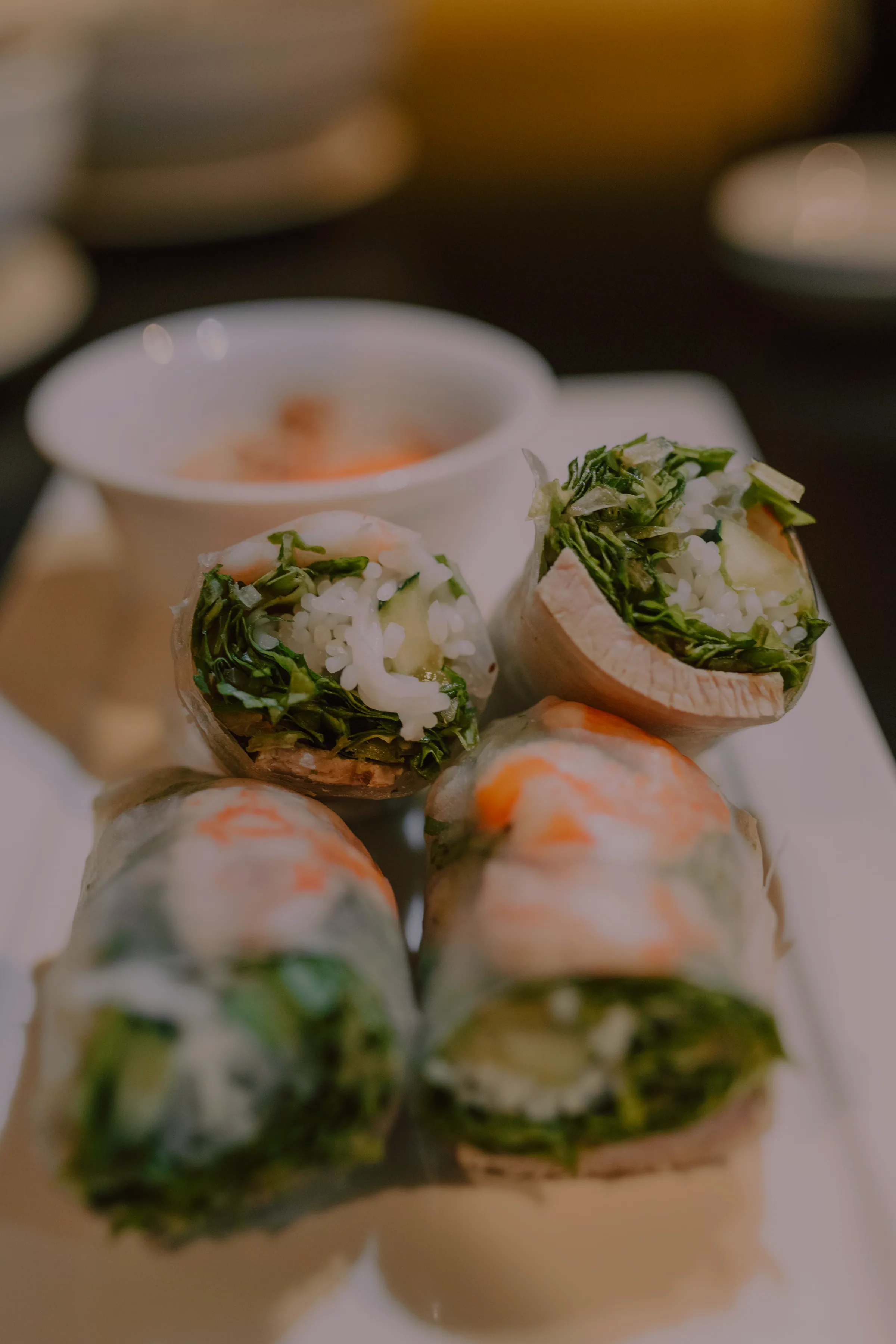 Four fresh summer rolls stood on end on a white plate, cut faces showing shrimp, rice, cucumber and shredded herbs, with a dipping sauce bowl blurred behind