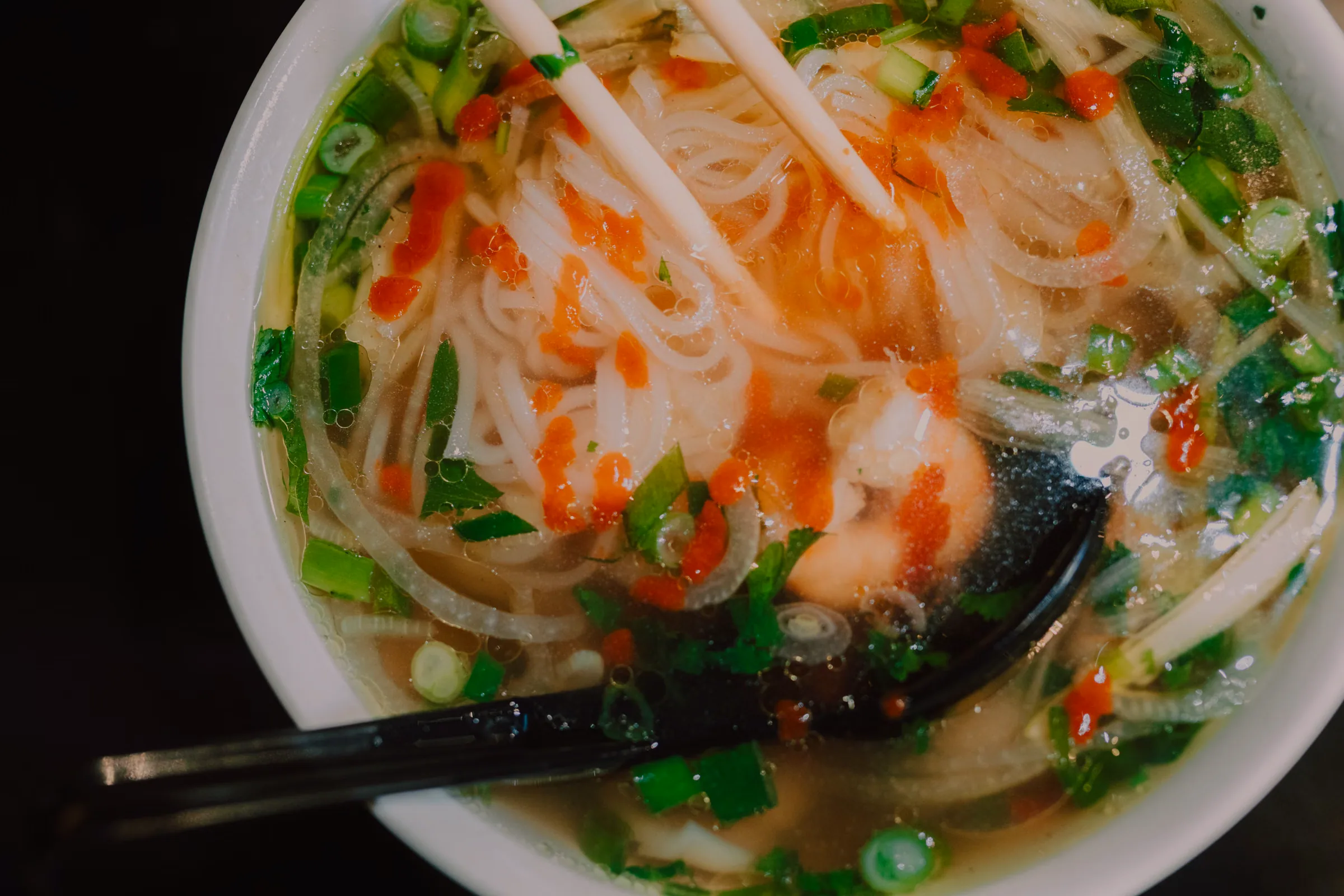 Overhead macro of a bowl of noodle soup: white rice noodles gripped by chopsticks, chilli sauce streaked across the surface, scallion and coriander floating on clear broth beaded with oil