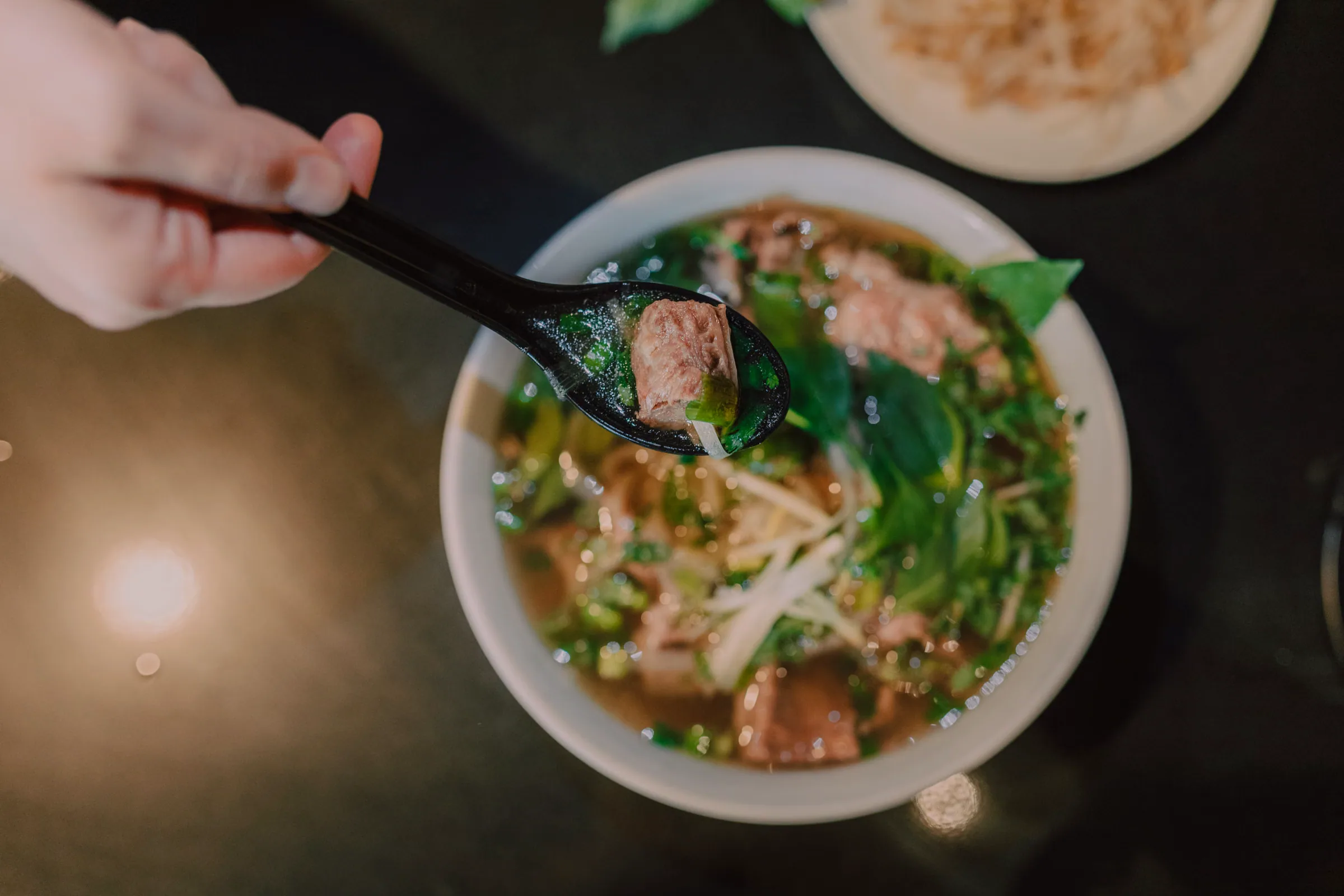 Seen from above, a hand holds a black soup spoon carrying a piece of beef and a basil leaf over a bowl of pho, with a side plate of bean sprouts at the top edge