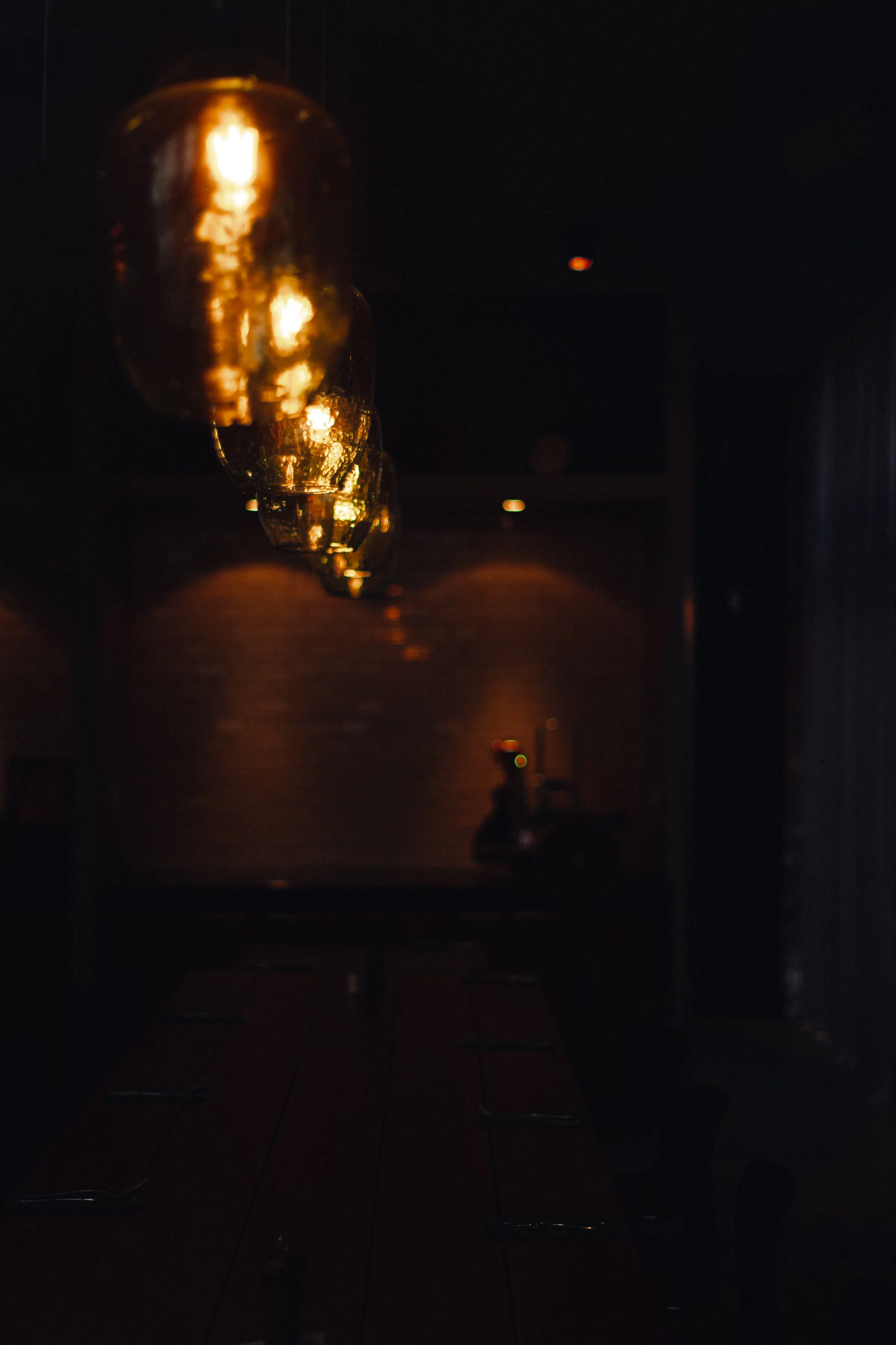 A dimly lit dining room with a row of glass pendant bulbs glowing warm amber in the foreground, a long communal table receding into darkness below a faint brick wall