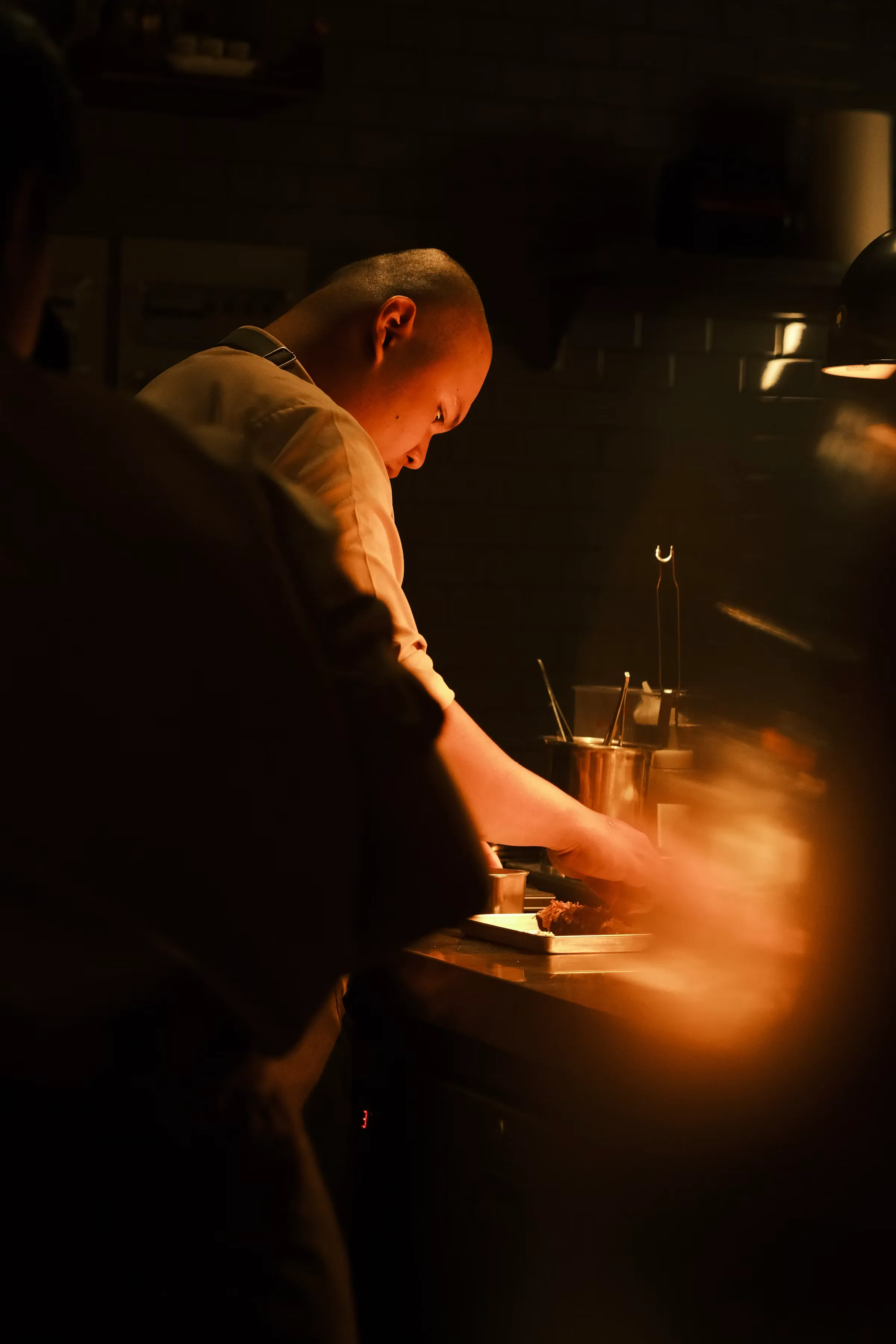 A cook in a pale apron leans over a stainless pass in a very dark kitchen, his face and arms lit by a single warm lamp, steam drifting through the light at the right of frame