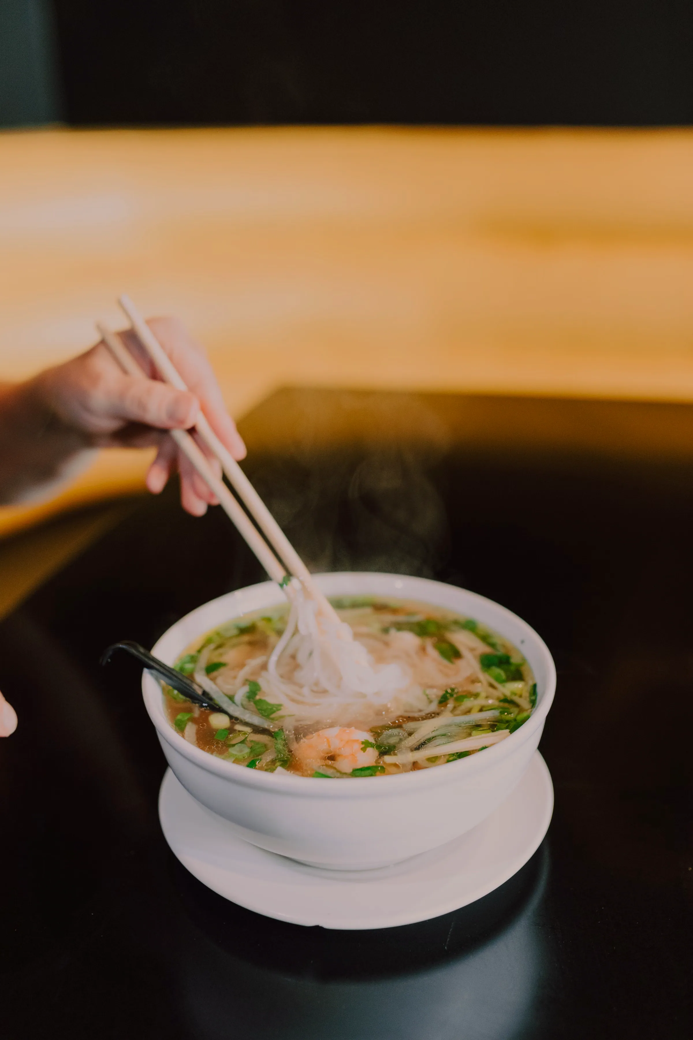 A hand lifts a tangle of rice noodles on pale wooden chopsticks out of a white bowl of pho, a plume of steam rising against a dark table and warm amber wall behind