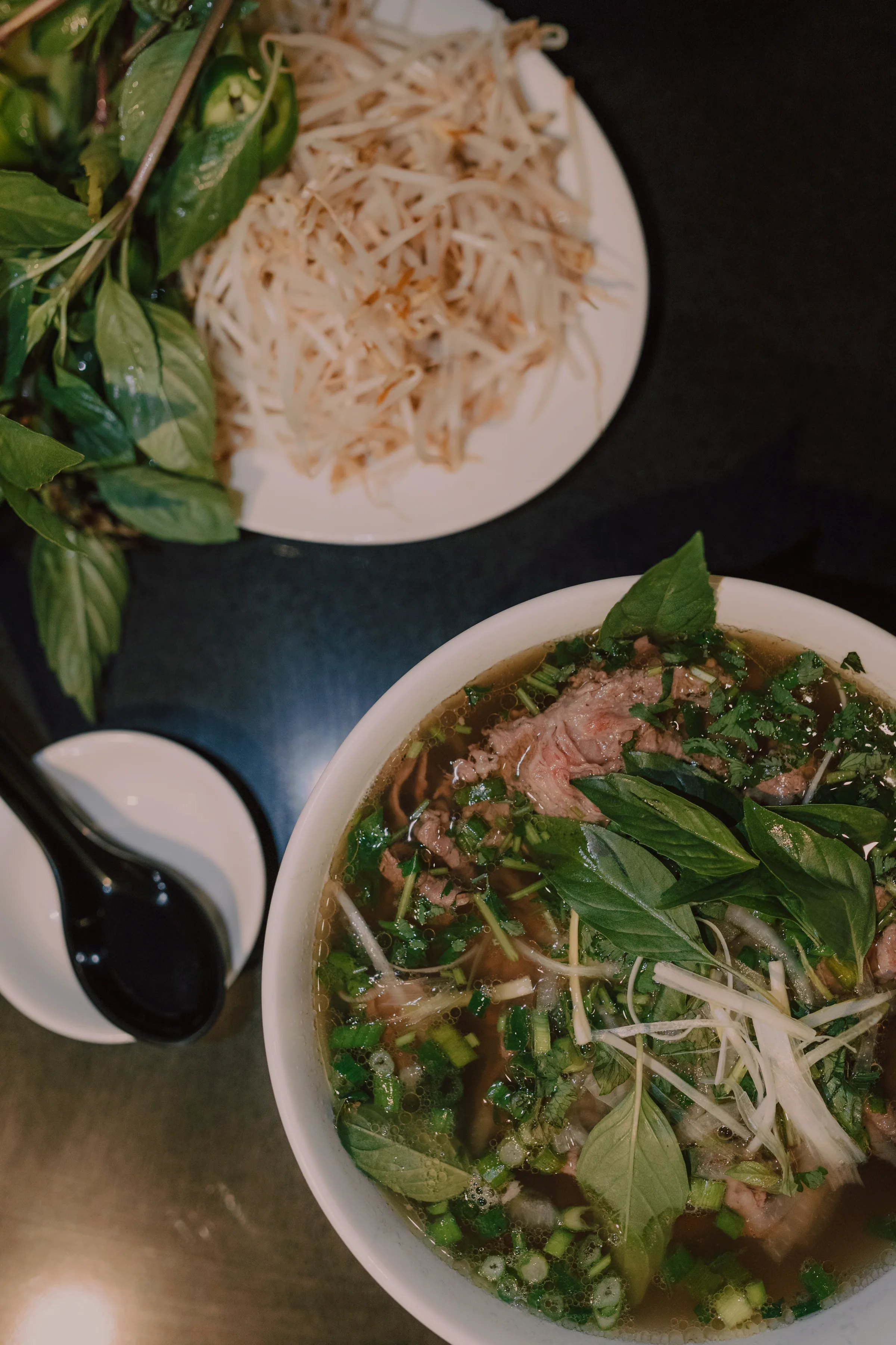 Overhead view of a side plate of Thai basil, bean sprouts and sliced green chilli next to a bowl of pho and a small dish of dark hoisin sauce on a dark grey table